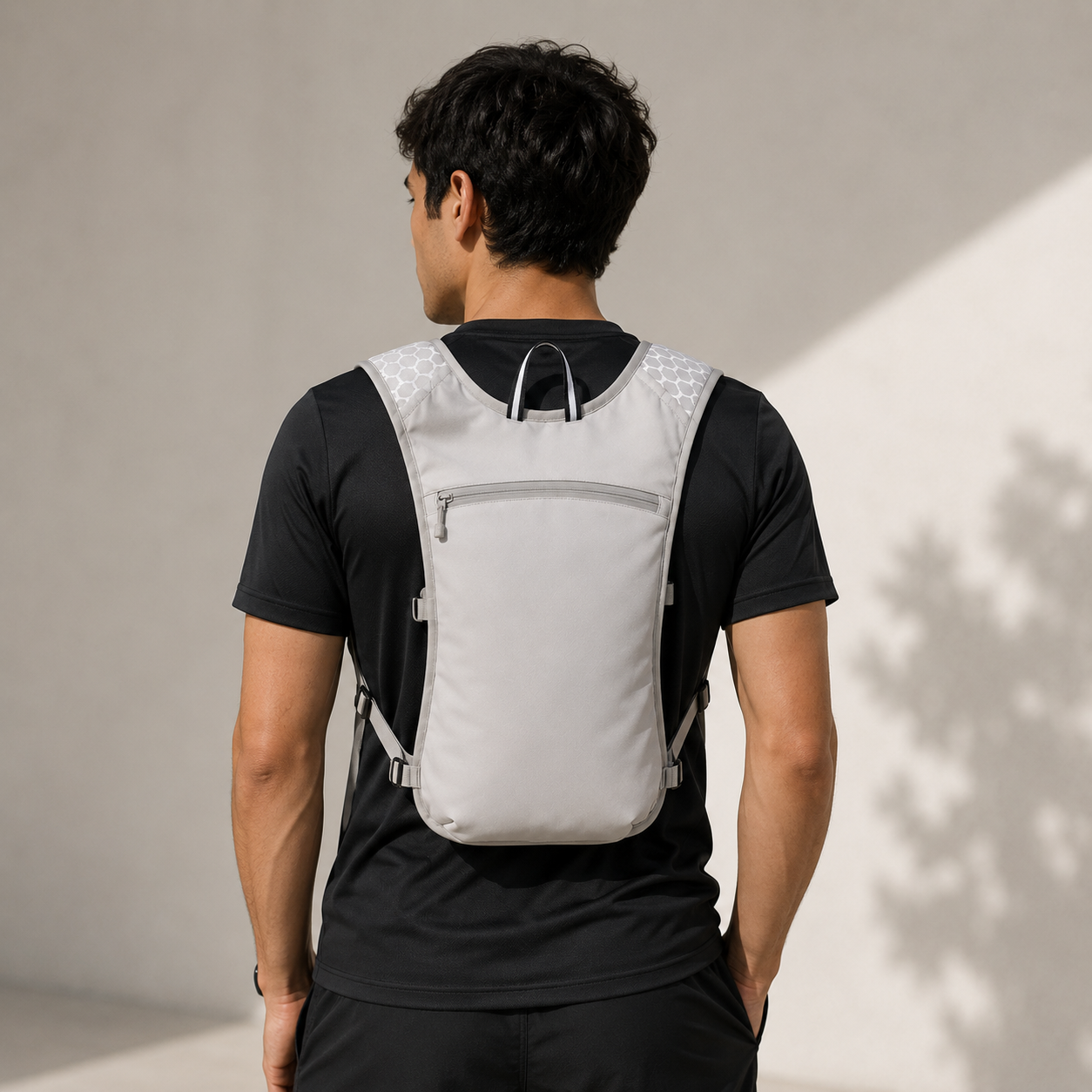 A man viewed from behind wearing a light gray and black hydration backpack with a front zippered pocket, set against a minimalist beige wall with soft shadows.