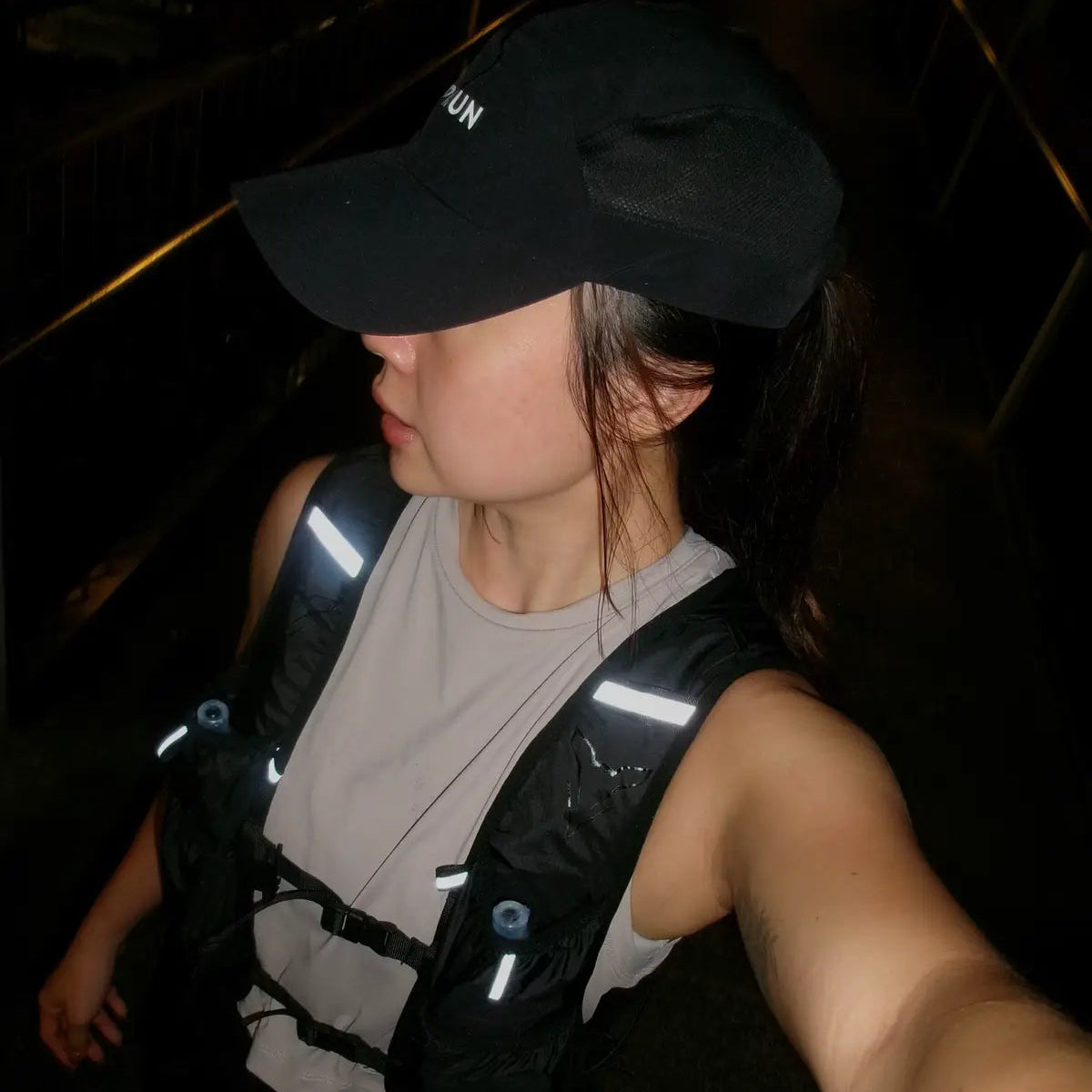A woman wearing a black baseball cap, a light gray sleeveless top, and a black hydration backpack with reflective white striping, captured in a dark, low-light selfie.