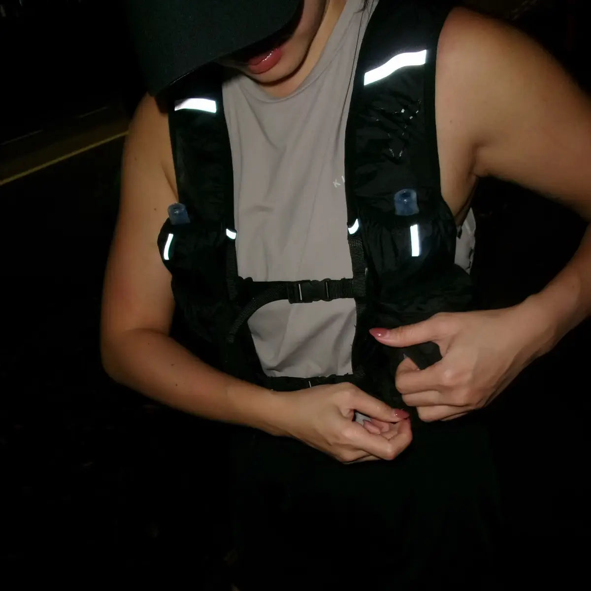 A person wearing a black reflective running vest over a light gray t-shirt and black running shorts, with a black cap, in a very dark setting.